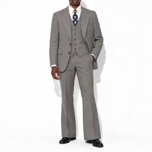 May include: A three-piece grey suit consisting of a single-breasted blazer, a matching button-up vest, and flared trousers. The ensemble is paired with a crisp white collared shirt and a dark blue patterned necktie, finished with black shoes.