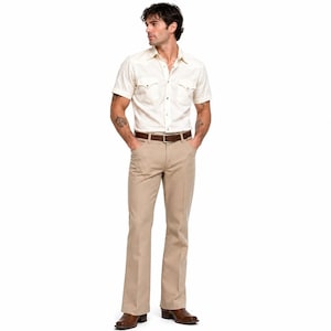 May include: A cream-colored short-sleeve shirt with button closures and a brown belt paired with tan flared pants. The outfit is completed with brown boots. The shirt has two front pockets and a classic collar.