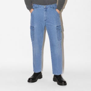 May include: Light blue denim cargo pants featuring a straight-leg cut, a button and zipper fly closure, belt loops, and two large rectangular side pockets on the thighs. The pants are paired with black leather boots and a dark grey long-sleeve shirt.
