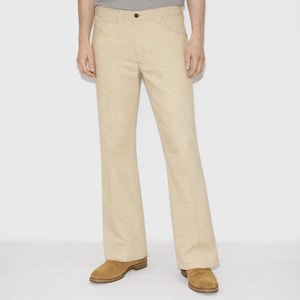 May include: Beige flared pants with a classic five-pocket design. The pants are made of a textured fabric and have a button and zipper closure. The pants are paired with brown suede shoes.