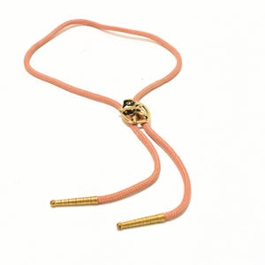 May include: A bolo tie with a peach-colored cord and gold-toned metal accents. The central piece features a detailed design, and the cord ends have decorative metal tips. The tie is set against a white background.