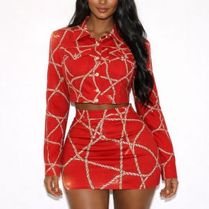 May include: A two-piece red outfit featuring a cropped jacket and a mini skirt. The set is adorned with a beige rope pattern. The jacket has long sleeves and button closures. The skirt is high-waisted.