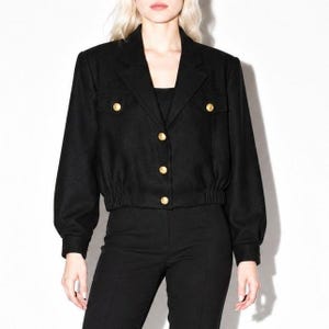 May include: A black cropped jacket featuring long sleeves, a collared neckline, and a button-down front. The jacket includes two chest pockets with flaps and gold-toned buttons, with an elasticized hemline at the waist for a fitted silhouette.