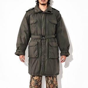 May include: An olive green long-length jacket featuring a high collar, a front zipper closure with a storm flap, two chest pockets, two large lower pockets, and a waist belt with a black plastic buckle. The sleeves have adjustable cuffs.