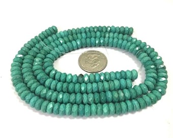 May include: A strand of faceted turquoise beads, coiled on a white surface. A US quarter is placed in the center for size reference. The beads are a vibrant teal color and have a polished finish.