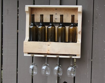May include: A wooden wine rack with five bottles of wine and four wine glasses hanging underneath. The rack is mounted on a gray wooden fence.