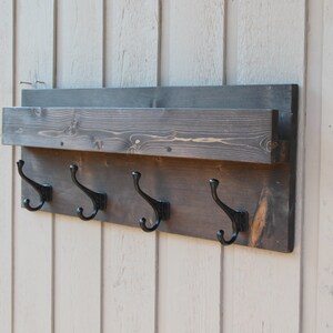 May include: A dark brown wooden wall mounted shelf with a small shelf on top and four black metal hooks underneath.