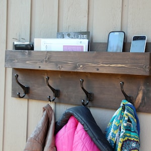 May include: A dark brown wooden coat rack with four black metal hooks and a shelf above. The shelf has two cell phones and some papers on it. There are three items hanging on the hooks, including a brown jacket, a pink jacket, and a colorful scarf.