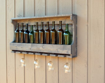 May include: A rustic wooden wine rack with a gray stain, holding eight wine bottles and six wine glasses. The rack is mounted on a light brown wall.