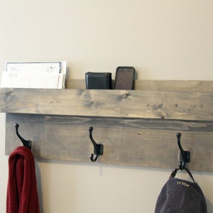 May include: A wooden wall-mounted organizer with a shelf and three black metal hooks. The shelf holds mail, a wallet, and a phone. A red scarf and a dark blue hat hang from the hooks. The wood is stained a gray color.