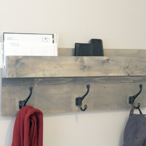 May include: A gray wooden wall-mounted organizer with a shelf and three black metal hooks. The shelf holds mail and a black object. A red scarf and a blue cap hang from the hooks. The wood has a distressed finish.