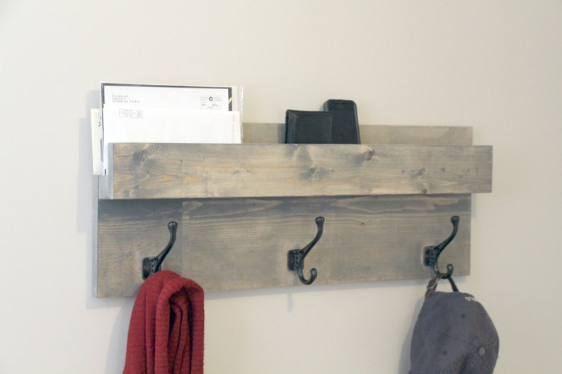 May include: A gray wooden wall-mounted organizer with a shelf and three black metal hooks. The shelf holds mail and a black object. A red scarf and a blue cap hang from the hooks. The wood has a distressed finish.
