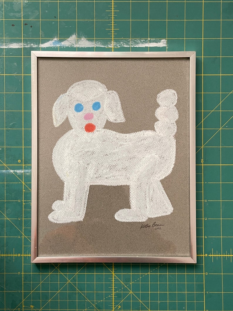 May include: A framed drawing of a white dog with blue eyes and a red nose on a brown background. The drawing is signed "Katie Bean 2021".