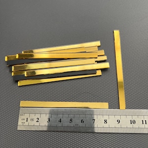 May include: A pile of 12 gold-colored metal strips, each measuring approximately 7.5 cm in length, with a black backing. A ruler is included for scale.