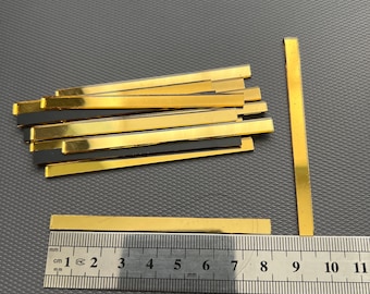 May include: A pile of 12 gold-colored metal strips, each measuring approximately 7.5 cm in length, with a black backing. A ruler is included for scale.