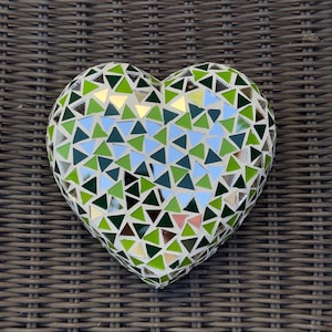May include: A heart-shaped mosaic sculpture featuring a variety of green, blue, and mirrored triangular tiles. The sculpture is set against a brown woven background. The mosaic tiles create a textured, decorative effect.