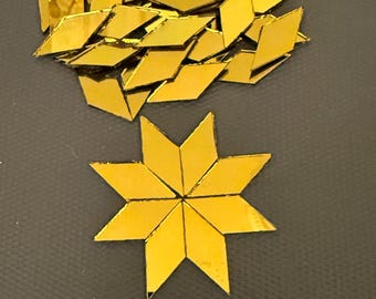 May include: A collection of small, gold-colored diamond-shaped mirror tiles. A cluster of loose pieces is arranged above a single eight-pointed star shape formed by eight of the diamond tiles joined together at their points.