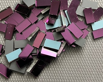Puede incluir: Un montón de pequeñas baldosas rectangulares iridiscentes en tonos de morado, gris y azul. Las baldosas están dispuestas en un patrón aleatorio y están dispersas sobre una superficie gris texturizada.