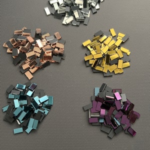 May include: Assortment of small, rectangular mirror tiles in various colours, including silver, gold, rose gold, teal, and purple. The tiles are arranged in five separate piles on a grey surface.