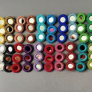 Puede incluir: Una colección de parches circulares bordados con espejos centrales rodeados de hilo en varios colores, incluyendo amarillo, marrón, azul, verde, rojo, morado, rosa y negro, dispuestos en un patrón de cuadrícula sobre una superficie gris texturizada.