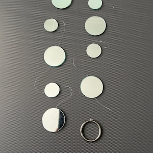 May include: An assortment of small, circular mirrors of different diameters, linked by fine, silver-coloured wires. A silver metal ring is positioned at the bottom. The mirrors are displayed on a textured, dark grey surface.