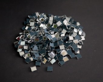 May include: A pile of small, square, silver-backed mirror tiles. The tiles are arranged in a random pattern and are scattered on a black surface.