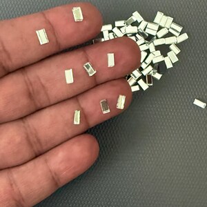 May include: A scattering of small, rectangular, silver-coloured beads. Some beads are scattered on a grey surface, while others are held in a hand. The beads have a reflective, metallic appearance.