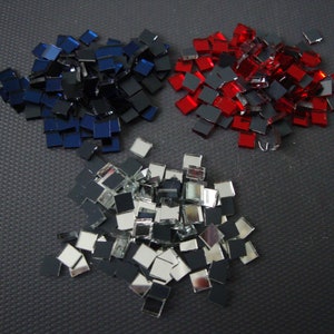 Könnte beinhalten: Ein Haufen kleiner quadratischer Spiegelkacheln in Rot, Blau und Silber. Die Kacheln sind auf einer schwarzen Oberfläche verstreut.