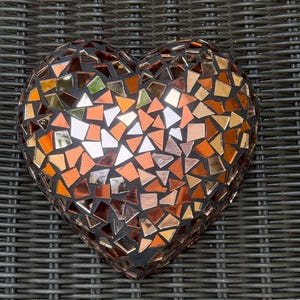 Peut inclure: Une mosaïque en forme de cœur composée de petites tuiles de verre irisées dans des tons de brun, orange et or. La mosaïque est placée sur un fond en osier gris foncé.