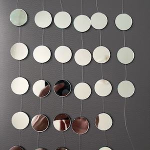 Puede incluir: Una hilera de azulejos redondos de espejo plateado con borde verde, colgando de una cuerda blanca. Los azulejos están dispuestos en filas de ocho, con un anillo en la parte inferior.