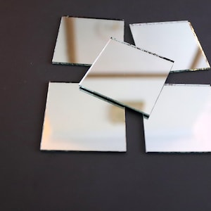 May include: Five small square mirrors with beveled edges are arranged in a pile on a dark gray surface.