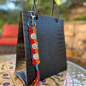 May include: A black leather tote bag with a silver key chain and a colorful macrame tassel. The tassel is made with red, orange, blue, and turquoise yarn and features silver beads.