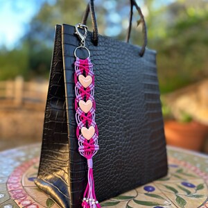 May include: A pink and purple heart-shaped keychain with a silver charm attached to a black crocodile embossed handbag.