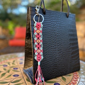 May include: A pink and blue macrame keychain with a silver ring and a silver clasp. The keychain has five colorful oval beads and a pink and white tassel.