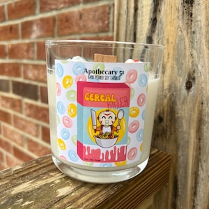 May include: A glass candle jar with a white label featuring a cartoon illustration of a person wearing a hockey mask holding a knife and spoon. The label reads "Apothecary 51 Hand Poured Soy Candles Cereal Killer 12oz | 80+ Hours". The candle is surrounded by colorful cereal rings.