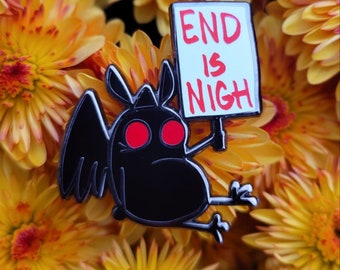 May include: A black enamel pin featuring a stylized bat-like creature with bright red eyes. The creature holds a white rectangular sign with the red text 'END IS NIGH' written in capital letters. The pin has a black metal outline.