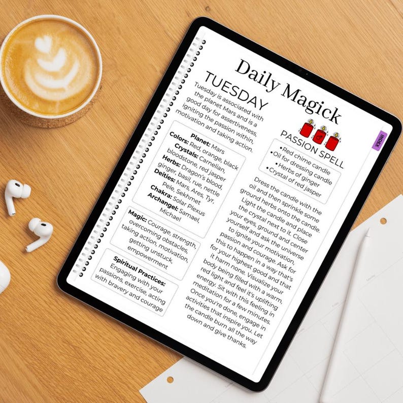 May include: A digital tablet displays a "Daily Magick" page with text about Tuesday, Mars, and a passion spell. The page includes a list of colors, crystals, herbs, deities, and a chakra. A cup of coffee and wireless earbuds are on the wooden surface.