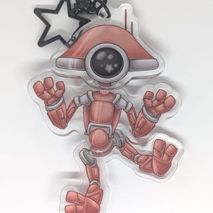 May include: A clear acrylic keychain featuring a cartoon robot in shades of red, gray, and white. The robot has a large, round eye and is posed with its arms and legs outstretched. A black star-shaped charm and a keyring are attached.