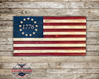Könnte beinhalten: Eine amerikanische Holzflagge mit 13 roten und weißen Streifen und einem blauen Kanton mit 13 weißen Sternen in einem Kreis mit dem Jahr 1776 in der Mitte. Die Flagge wird vor einem Holzmaserungshintergrund gezeigt.