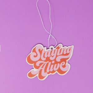 May include: A pink and orange car air freshener with the text "Staying Alive" in a retro font.