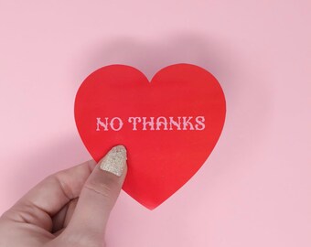 Op de afbeelding: Een rood papieren hart met de tekst "NO THANKS" in wit erop. Het hart wordt vastgehouden door een hand met een gouden glitternagel.