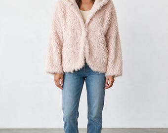 Puede incluir: Una chaqueta de piel sintética de color rosa pálido con capucha, mangas largas y frente abierto, combinada con una camiseta color crema, pantalones vaqueros azules y zapatillas blancas. La chaqueta tiene una textura peluda.