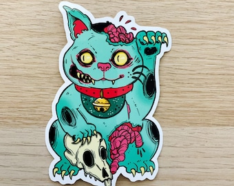 Puede incluir: Una figurita de gato zombie verde turquesa que sostiene una calavera y cerebros en sus patas. El gato tiene un collar rojo con una campana dorada y ojos amarillos. La figurita es una parodie de un Maneki-Neko japonés tradicional, o "gato de la suerte", amuleto de la buena suerte.