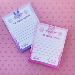 May include: Two pink and purple gradient notepad sets with a cartoon devil illustration and the text "Soul Binding Contract". The bottom notepad has the signature line "Seitan CEO of Hell".