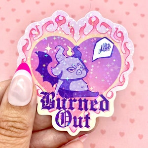 May include: A holographic sticker with a purple cartoon demon with horns and wings. The demon is sitting in a pink heart with flames around it. The text "Burned Out" is written below the heart.
