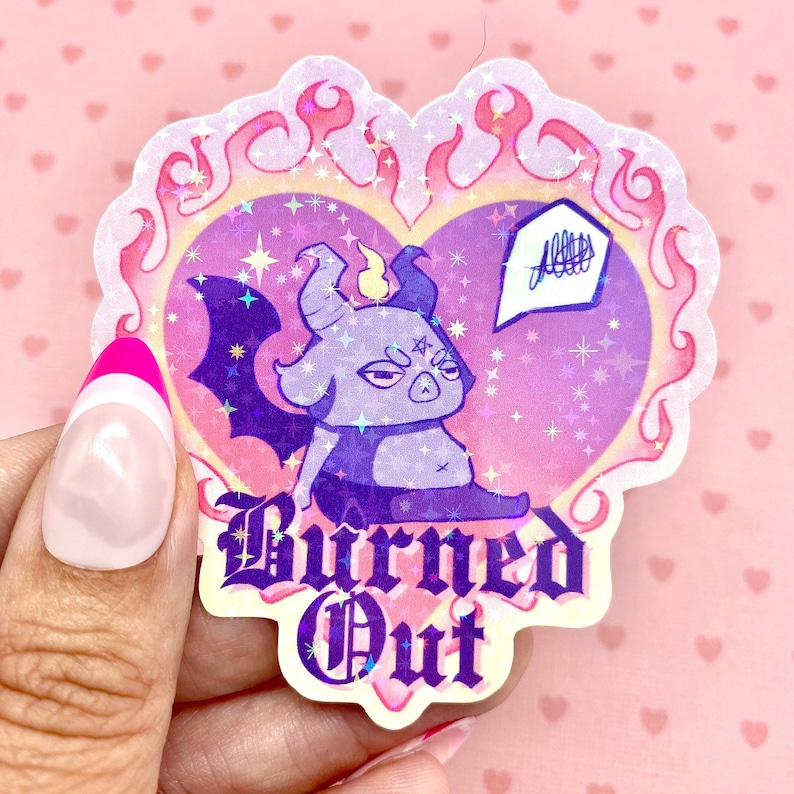 May include: A holographic sticker with a purple cartoon demon with horns and wings. The demon is sitting in a pink heart with flames around it. The text "Burned Out" is written below the heart.