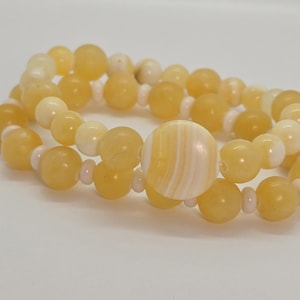 May include: A double-strand bracelet featuring round yellow and cream-colored beads. The design alternates between translucent yellow spheres and smaller, opaque cream-colored spacers, centered with a large, flat, circular bead with white and yellow stripes.