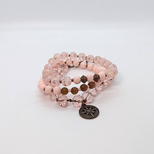 May include: A stack of three wrap bracelets. The top and bottom bracelets are made of faceted pink beads. The middle bracelet has pink and brown beads. A bronze compass charm hangs from the bottom bracelet.