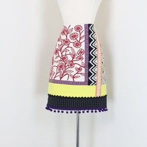 May include: A colorful, patterned skirt displayed on a mannequin. The skirt features a floral design in red, a yellow scalloped hem, and a black and white checkered bottom with purple pom-poms. The skirt is on a chrome stand.