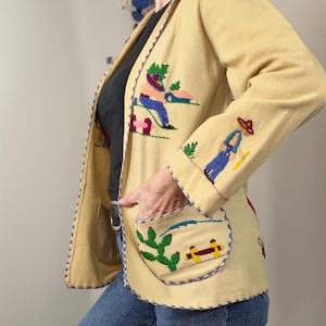 May include: A light yellow jacket featuring colorful embroidered scenes of figures in hats and cacti. The garment has long sleeves, a front pocket, and decorative blue cross-stitch detailing along the edges, worn over a black top and blue jeans.